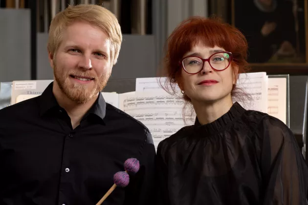 Albina Veisland och Pietari Huhtinen står framför orgelns notställ de tittar in i kameran. Båda har svarta kläder.